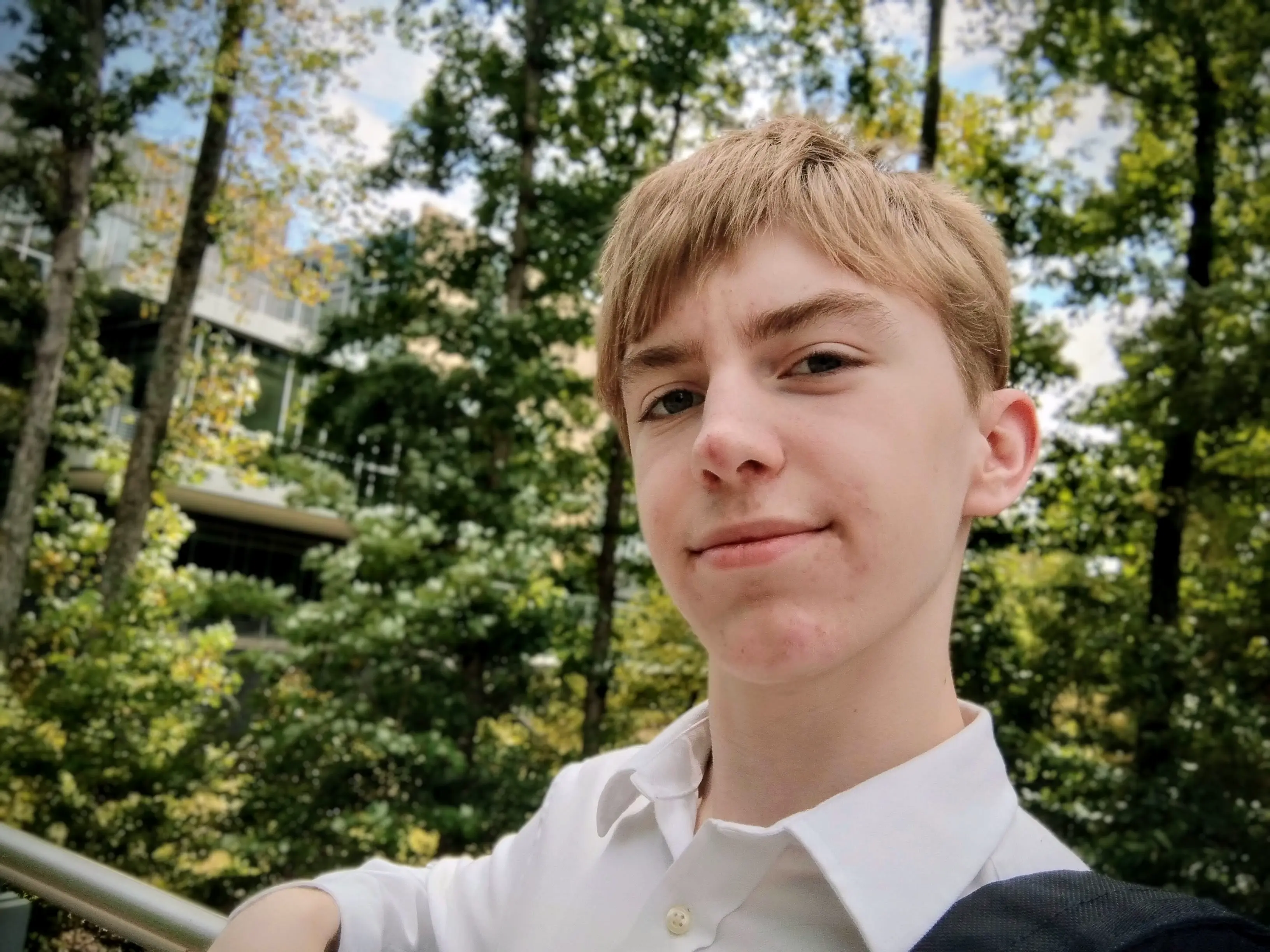 Ethan, a fair-skinned brown-haired teenager, wearing a white-collared button-up shirt, gently smiling at the camera while leaning against a railing. A vibrant, slightly blurred forest is visible in the background Ethan, a fair-skinned brown-haired teenager, wearing a white-collared button-up shirt, gently smiling at the camera while leaning against a railing. A vibrant, slightly blurred forest is visible in the background