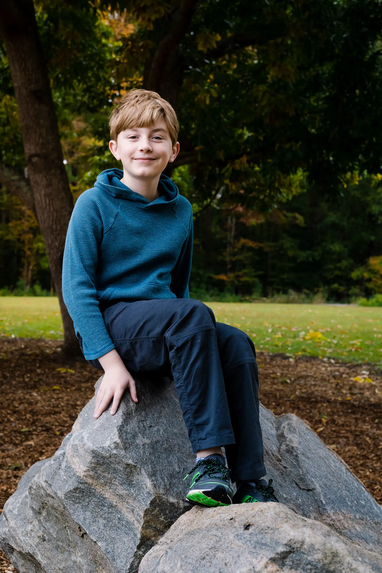 Ethan, a fair-skinned brown-haired teenager, sitting on a boulder and smiling pleasantly underneath a tree
